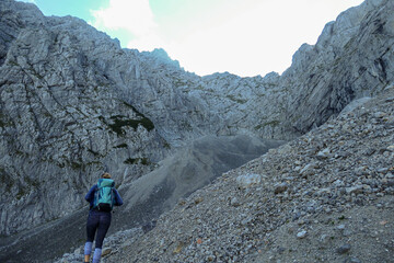 A woman with backpack hiking through steep and high mountain crater in Austrian Alp. The sun shines behind the mountain wall. The whole area is covered with shadow. Steep and narrow path along.
