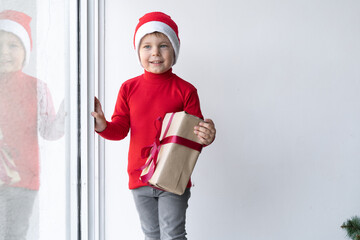 a little boy dressed in a santa claus hat stands near the window holding a box with a gift.