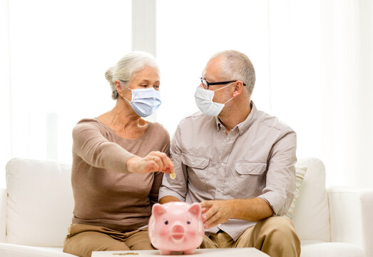 savings, health and old age concept - senior couple wearing face protective medical mask for protection from virus disease putting coin into big piggy bank