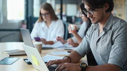 Man in eyewear and formal clothes working by using laptop in the office. Colleagues at background.
