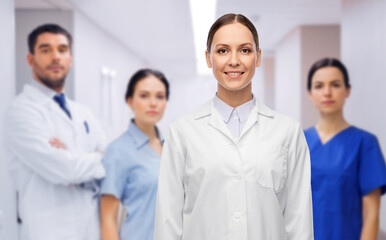 medicine, profession and healthcare concept - happy smiling female doctor in white coat over group of colleagues at hospital on background