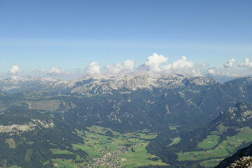 A panoramic view on a distant chain of Alps from another high peak. The massive chain is partially shrouded with clouds. Below there is lush green valley and a few cities in it. Austrian landscape