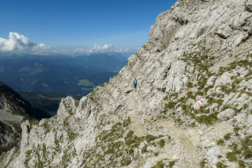 Woman walking on a very narrow and steep pathway along a mountain wall in Austrian Alp, leading to the top of Grimming. The path is marked with red-white-red sign. Dangerous trail. Following the path