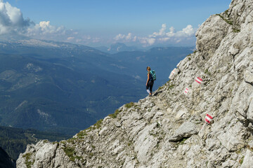 Fototapeta na wymiar Woman walking on a very narrow and steep pathway along a mountain wall in Austrian Alp, leading to the top of Grimming. The path is marked with red-white-red sign. Dangerous trail. Following the path