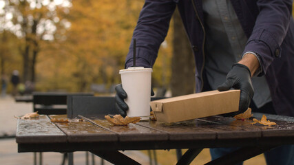 Close up male hands in a black protective gloves placing food box and a plastic cup from fast food restaurant on the table outdoors. Yellow leaves and water drops on the table. Autumn day in the city
