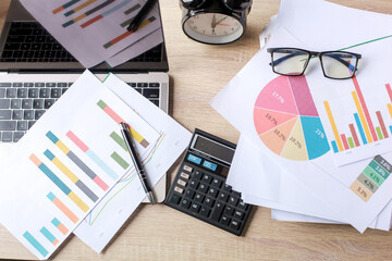 Flat lay top view of Messy office desk. Business accessories on wooden office desk: laptop, glasses, pen, charts on white sheets, calculator and alarm clock