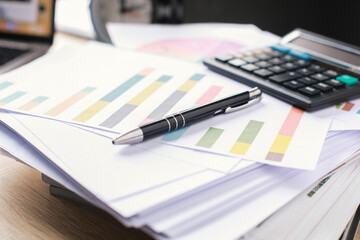 office desk with laptop, calculator, pen, stack of business reports and alarm clock. Pen on charts on white sheets with blurry on the background