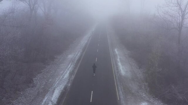 young sporty man in tracksuit clothes runs along a flat asphalt road along white markings surrounded by trees in frost in winter in foggy weather, 4K aerial video from drone