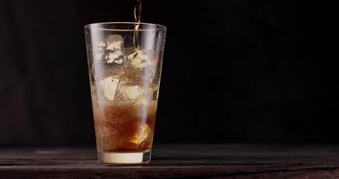 Cola is poured into a glass of cola with ice. The glass stands on a wooden table with a black background. Gas bubbles actively rise in the glass. Slow motion, Blackmagic Ursa Pro G2.