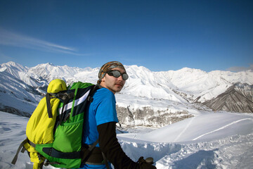 Man in bright sportive clothes and sun glasses with green backpack hiking in snowy mountains in Georgia. Copy space. Solo traveller and winter escape concept