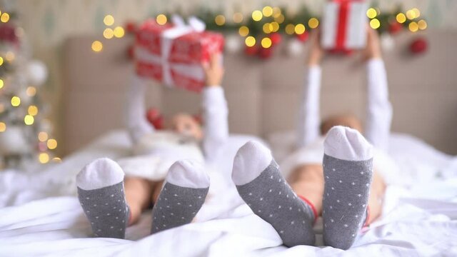 Close up kids feet lying on bed at home in christmas decorated room. Girls hold their gifts with lights on background
