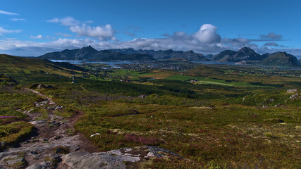 Beautiful panorama view over the west of Vestvågøy island with rocky hiking trail, green vegetation, antenna tower as well as Leknes town and rugged mountains in background sunny day in late summer.