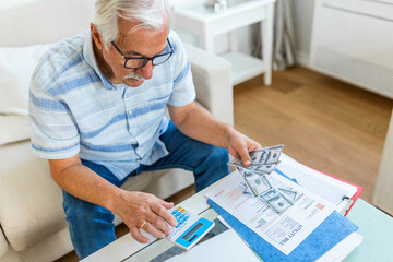 Serious elder senior grey-haired grandpa using calculator counting bank loan payment holding papers at home, focused mature old man doing paperwork calculating bills managing finances sit on sofa