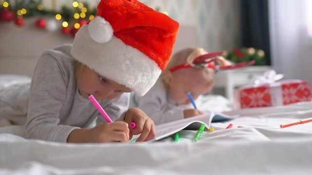 Two cute little girls sisters write letter to Santa Claus lying on bed at home in Christmas decorated room