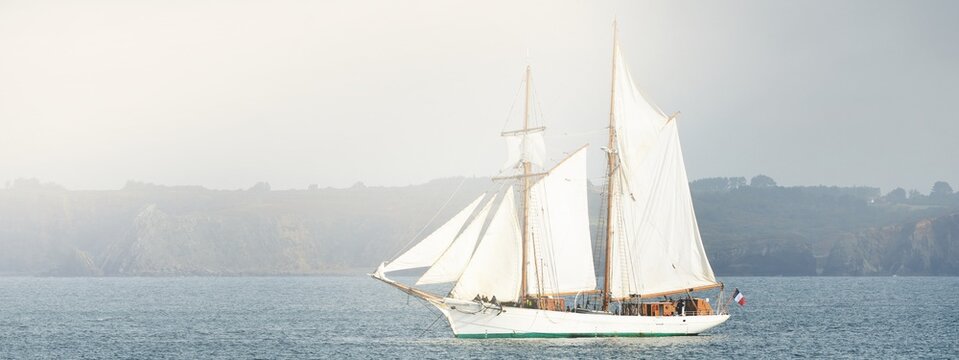 An elegant two-masted gaff schooner (tall ship) near the rocky shores of Brittany, France. Travel, history, transportation, sailing, sport, cruise, regatta, nautical vessel. Panoramic view
