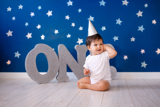 Baby boy of one year old wearing a white body and a festive paper hat sits on the floor on a blue background with stars and big silver letters