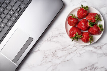 Open laptop computer and red strawberries in plate on table, copy space. Freelancer workspace, home...