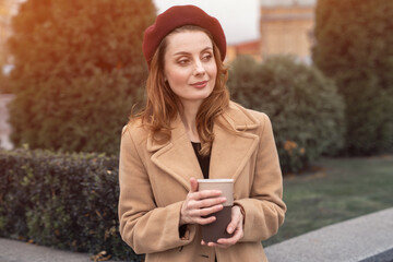 Pensive beautiful young woman holding a cup of coffee on the street. Female fashion concept. Portrait of stylish young woman wearing autumn coat and red beret outdoors. Tinted photo. 