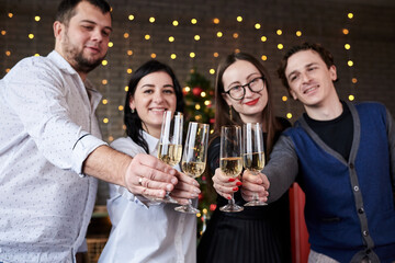 New year party celebration with friends and family at home. Four young people, wearing casual clothes, holding champagne glasses in dark room with christmas lights, smiling, laughing, doing cheers.
