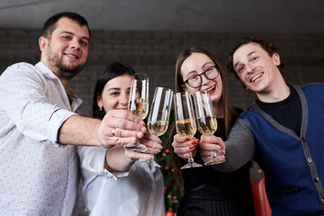 New year party celebration with friends and family at home. Close-up picture of four young people, wearing casual clothes, holding champagne glasses in dark room, smiling, laughing, doing cheers.