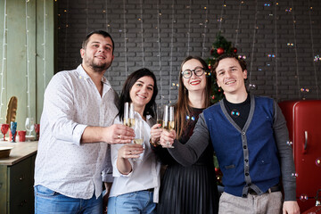 New year party celebration with friends and family at home. Four young people, wearing casual clothes, holding champagne glasses in dark room with christmas lights, smiling, laughing, doing cheers.