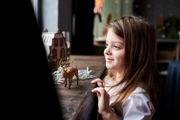 Small girl, standing by windowsill, playing with wooden toy house and deer in dark room with christmas decorations. Winter holidays at home. Leisure activity for children during quarantine time.