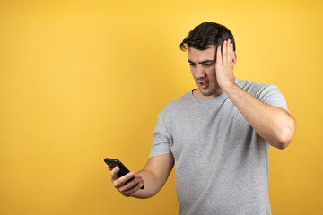 Young handsome man wearing a casual t-shirt over isolated yellow background holding a phone with surprised expression