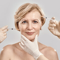 Close up portrait of beautiful naked middle aged woman looking at camera while she gets injections in her face, posing isolated against grey background