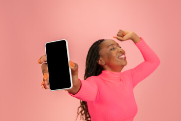 Naklejka premium Shows phone screen. Happy young beautifil african-american woman isolated on pink background. Copy space for ad, design. Half-length shot. Concept of human emotions, facial expression.