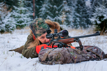 A beautiful young hunter woman in camouflage lies on the ground and looks into the rifle. Winter...