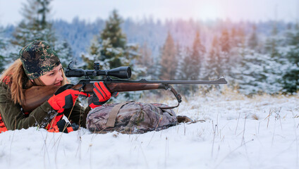 A beautiful young hunter woman in camouflage lies on the ground and looks into the rifle. Winter...