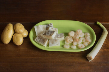 A wooden table with potatoes, leek, and a green platter with cod fillets and prawns. Ingredients for cooking fish and vegetables.