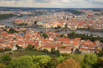 Fototapeta na wymiar Cityscape of Prague, Czech Republic. View from a hill to the orange tiled roofs and light walls of houses. Green forest in the foreground, river with numerous bridges in the center of the frame.
