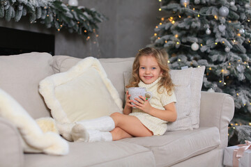 A white girl with a charming smile in a knitted white dress and socks with rings on her hands holds a cup in her hands sits on a soft sofa with pillows against the background of a Christmas tree