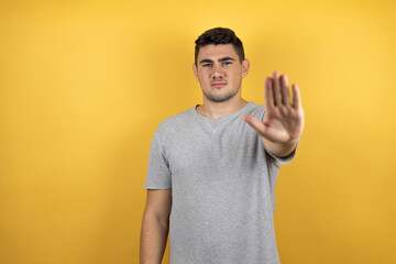 Young handsome man wearing a casual t-shirt over isolated yellow background serious and doing stop sing with palm of the hand.