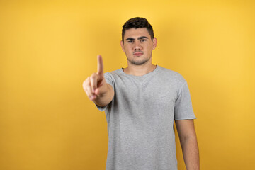 Young handsome man wearing a casual t-shirt over isolated yellow background showing and pointing up with fingers number one while is serious