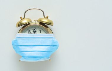 Vintage alarm clock partially covered by a medical mask on a white background. Concept - a pandemic...