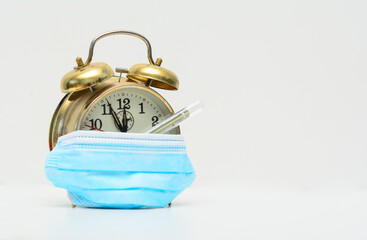 Vintage alarm clock partially covered by a medical mask on a white background. Under the mask is a...