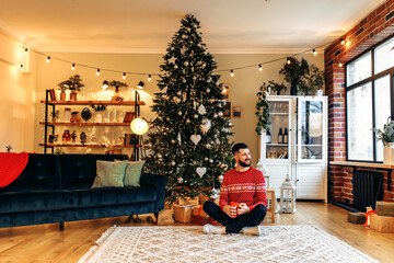 Handsome young man in a Christmas sweater, drinking coffee or tea, sitting at home near the Christmas tree