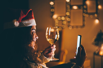 Happy young woman wearing Santa hat, drinking champagne, talking with friends on video call celebrate New Year party.
