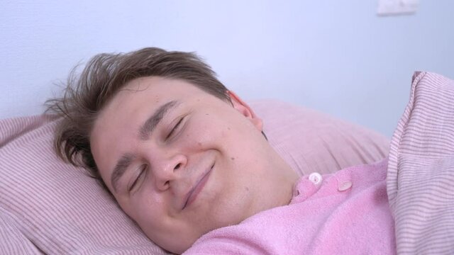Young caucasian man with somnambulism in pink pajamas sleeps sweetly at home, mumbles in his sleep and smiles because he sees pleasant dream, close up.