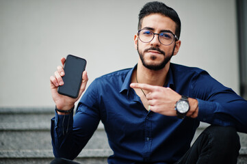 Middle eastern entrepreneur wear blue shirt, eyeglasses against office building sitting on stairs...