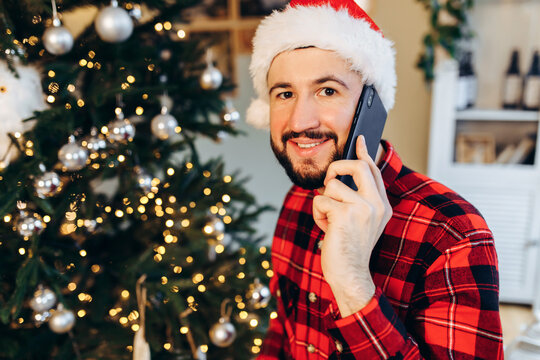 young man in Santa Claus hat talking on cell phone at home against background of Christmas tree