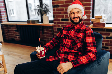 smiling man in Santa Claus hat with a cup of coffee sits on the sofa at home near the Christmas tree, Merry Christmas