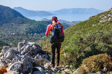 A man with a large backpack walks along a narrow rocky path overlooking the Mediterranean Sea....