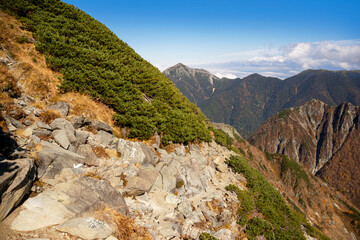 秋の紅葉の北アルプス、北穂高岳と涸沢をつなぐ登山道とペンキで道しるべの書き込みのある岩壁、ハイマツの茂み、遠くの常念岳と山々、青空、白い雲。
