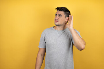 Young handsome man wearing a casual t-shirt over isolated yellow background surprised with hand over ear listening an hearing to rumor or gossip