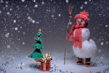 A snowman in a hat and scarf  looking at the tree and a gifted box. Smooth Black gradient background. Snowfall. Free space to text.
