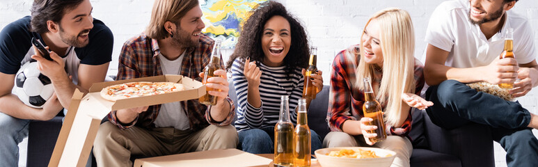 laughing african american woman pointing with finger while watching football competition with excited multiethnic friends, banner