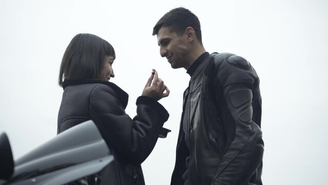 Happy Caucasian girlfriend feeding Middle Eastern boyfriend with sweet tasty cookie outdoors. Smiling interracial couple of bikers in leather jackets enjoying cloudy day. Dating and romance.